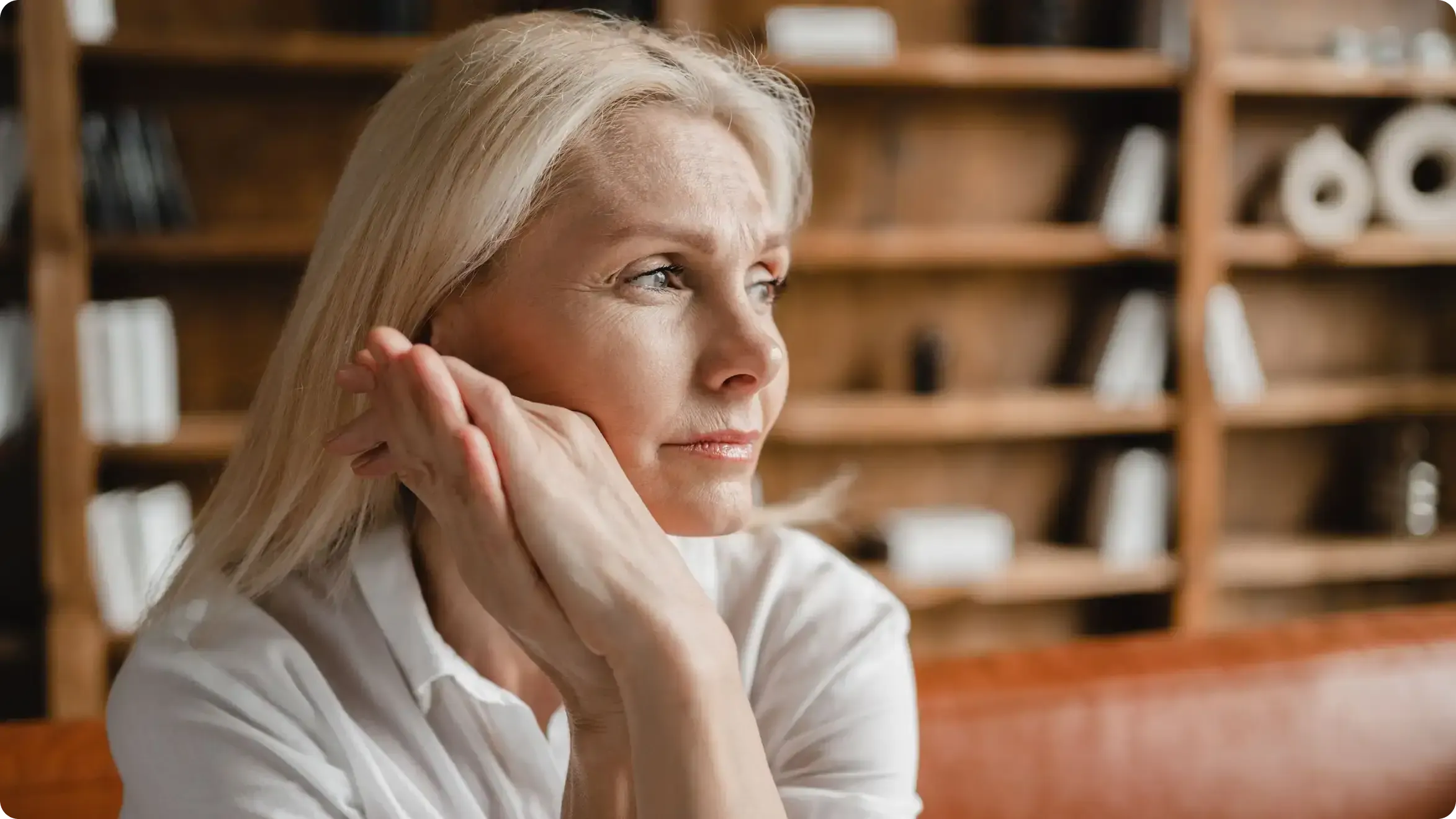 Middle-aged woman sitting on a leather couch, looking thoughtful and gazing into the distance with hands gently resting near her face.